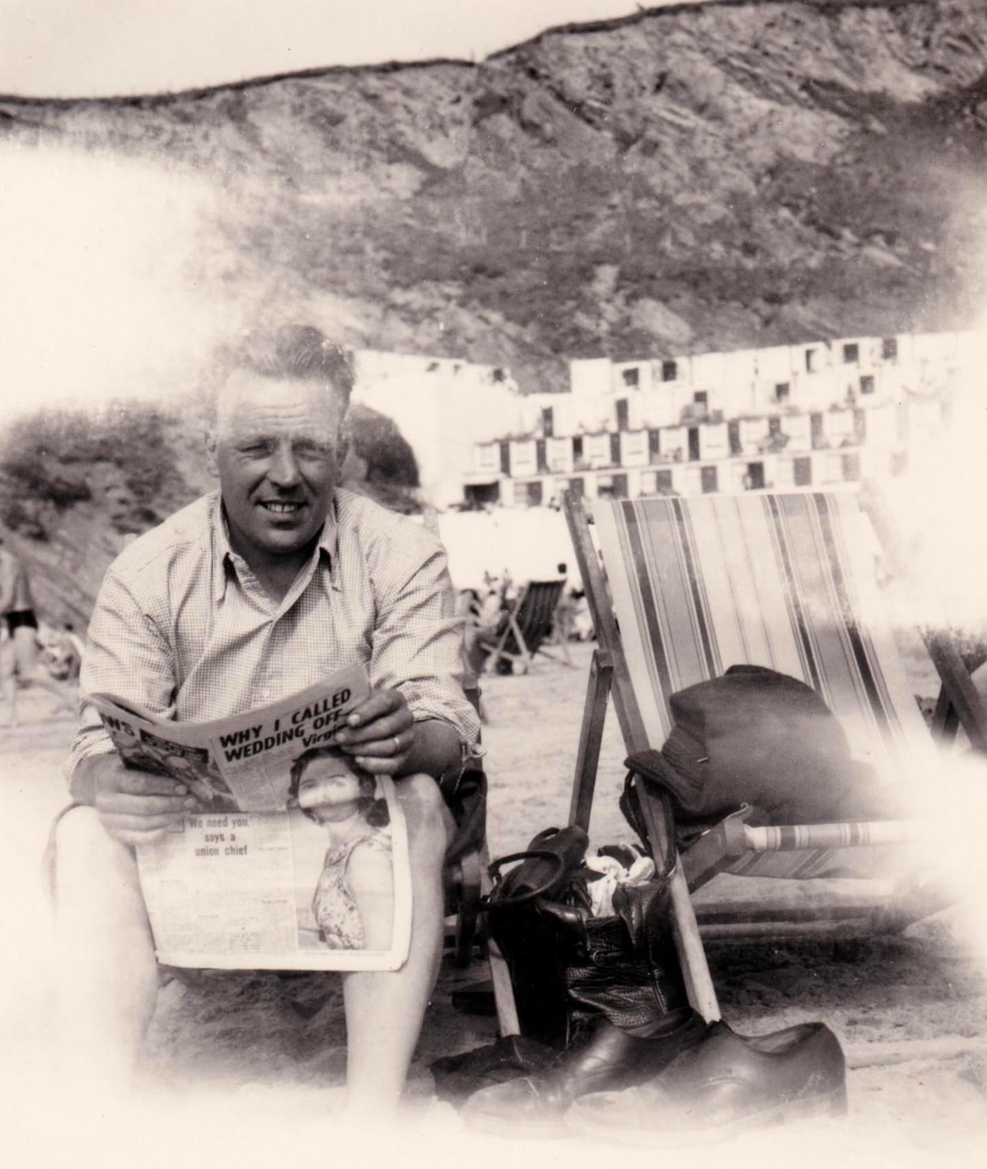 Jack is sitting on one of two deckchairs on a beach, leaning forward in the chair and smiling for the camera. He holds a newspaper where a headline, "Why I Called Wedding Off", clearly visible.