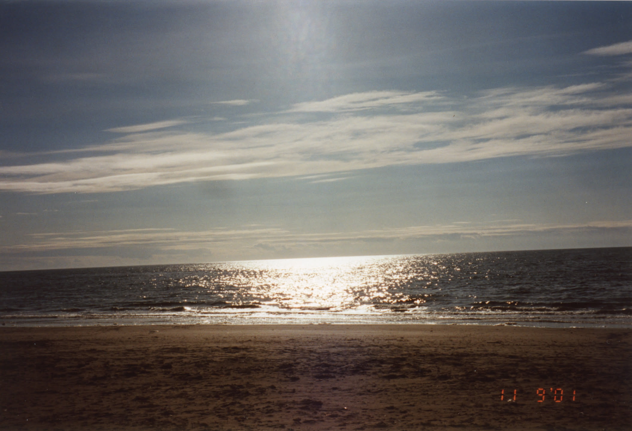 A view towards a sunset over the beach at Ayr. The photo isn't level, so the sea is at a slight slope going up towards the right. The sky is blue with tinges of orange and light, wispy clouds. Sunlight reflects off the rippling sea. A timestamp in the bottom-right corner reads 11-9-01: 11th September 2001.