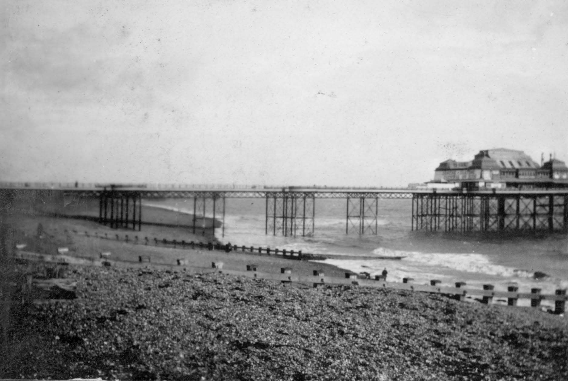 A black and white photo of Folkestone Pier with the beach and groynes in the foreground. The pier itself is intact and there appears to be some people walking on the pier.