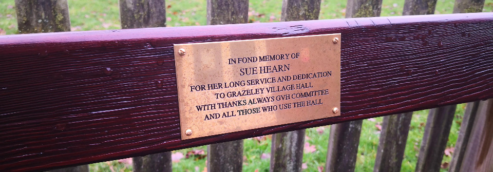 A memorial plaque on a bench. It reads In fond memory of Sue Hearn for her long service and dedication to Grazeley Village Hall with thanks always GVH committee and all those who use the hall