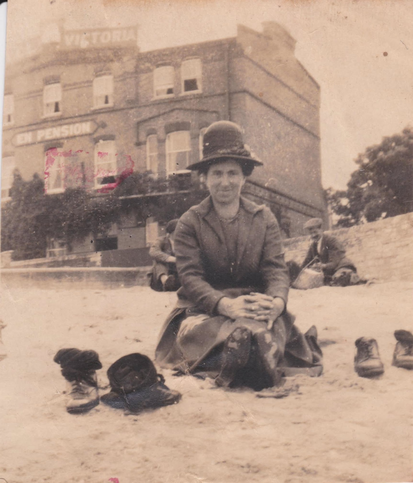 A photo of a woman, dressed in a long skirt and a long coat, with a tall cloche hat. In the background is a building with a sign that reads "Hotel Victoria" - "En pension".