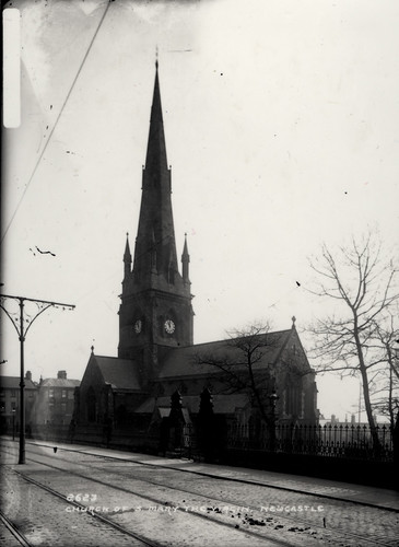 A black and white photo of a church with a tall spire and a clock on the two visible bell tower walls. Tram lines run in the road in front of the church.